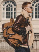 Man_wearing_a_brown_leather_jacket_with_fur_lining_holding_a_matching_backpack_in_front_of_a_building.-1